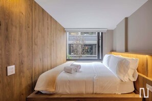A modern bedroom with a wooden headboard, a large window with a view of the outside, and a neatly made bed with white linens. - NOK Amazing 1BR Apartment With Natural Light (Bogotá)
