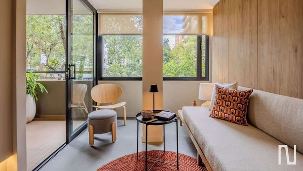 Habitación de hotel moderna con sofá, una silla, una mesita y un ventanal grande que ofrece vistas a árboles y edificios.