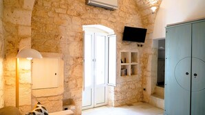 A stone-walled room with a white door, a television mounted on the wall, a cabinet, and a lamp. - Dimora Dolce Vista - Arcuève (Ostuni)
