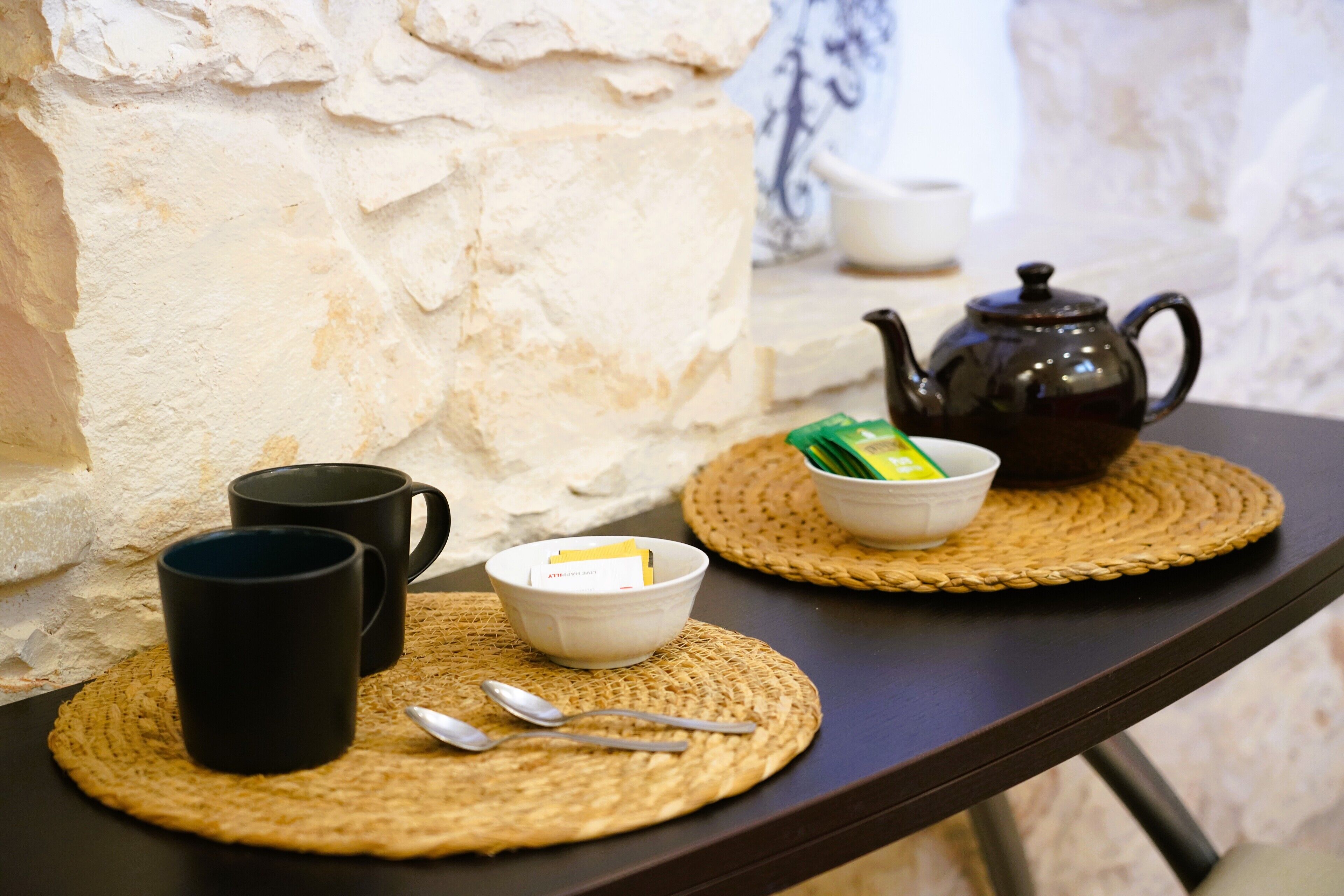 A table with two black mugs, a white bowl with a card, a teapot, and a box of tea on woven placemats.