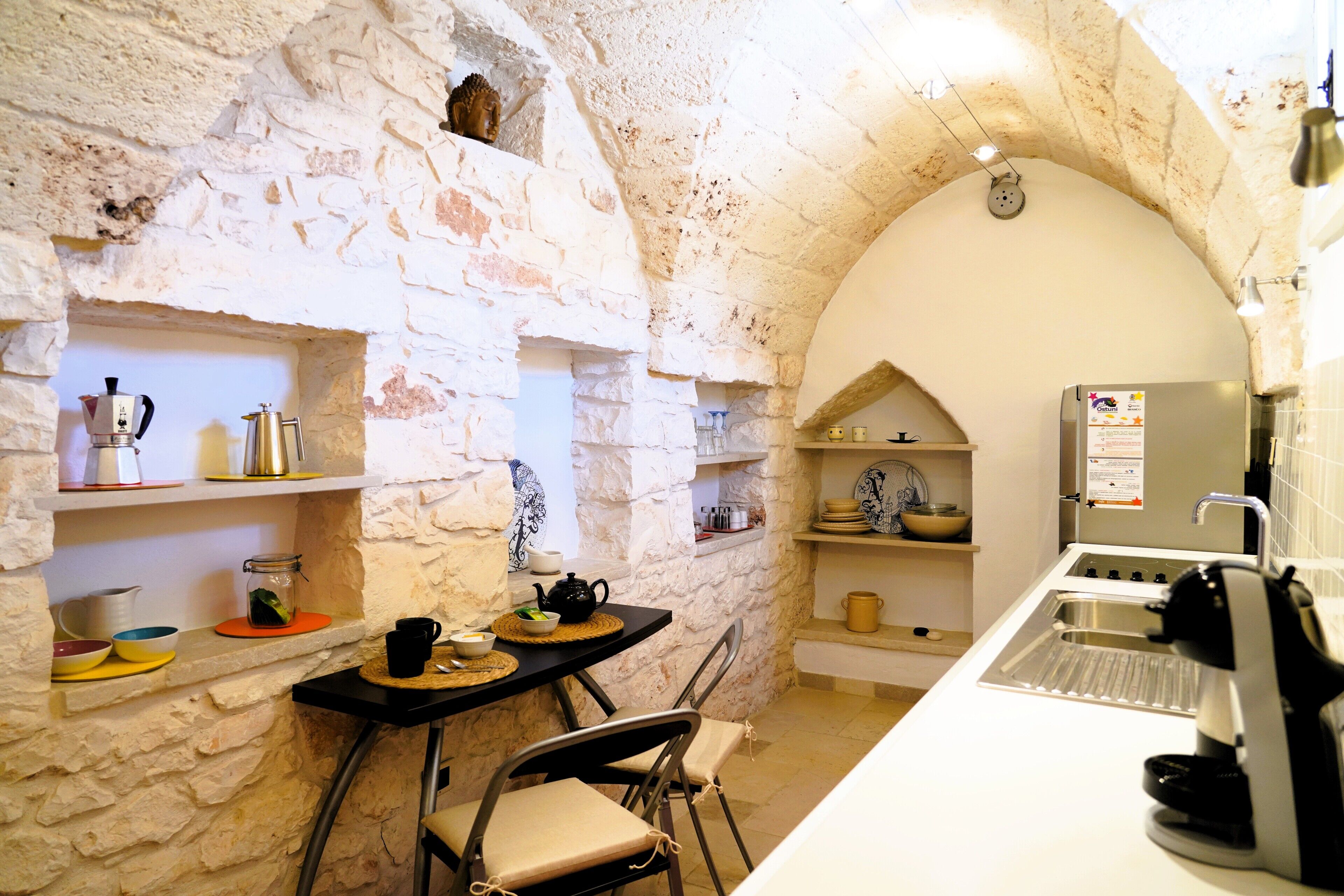 A compact kitchen with stone walls, a sink, a coffee machine, and shelves with dishes and a teapot.
