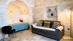 A room with a bed, a sofa, a chair, and a shelf with decorative items. - Dimora Dolce Vista - Arcuève (Ostuni)