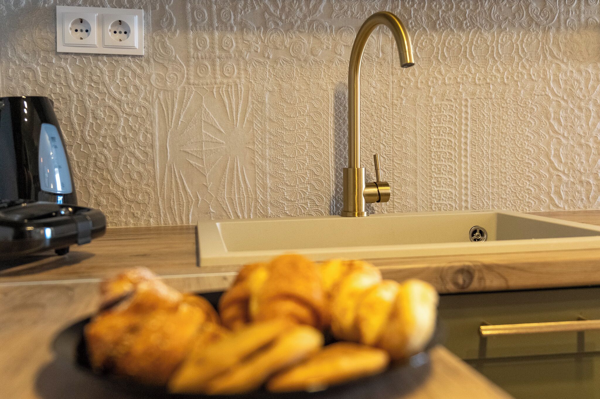 Une cuisine équipée d’un robinet moderne, d’une prise électrique et d’une assiette de croissants posée sur le comptoir.