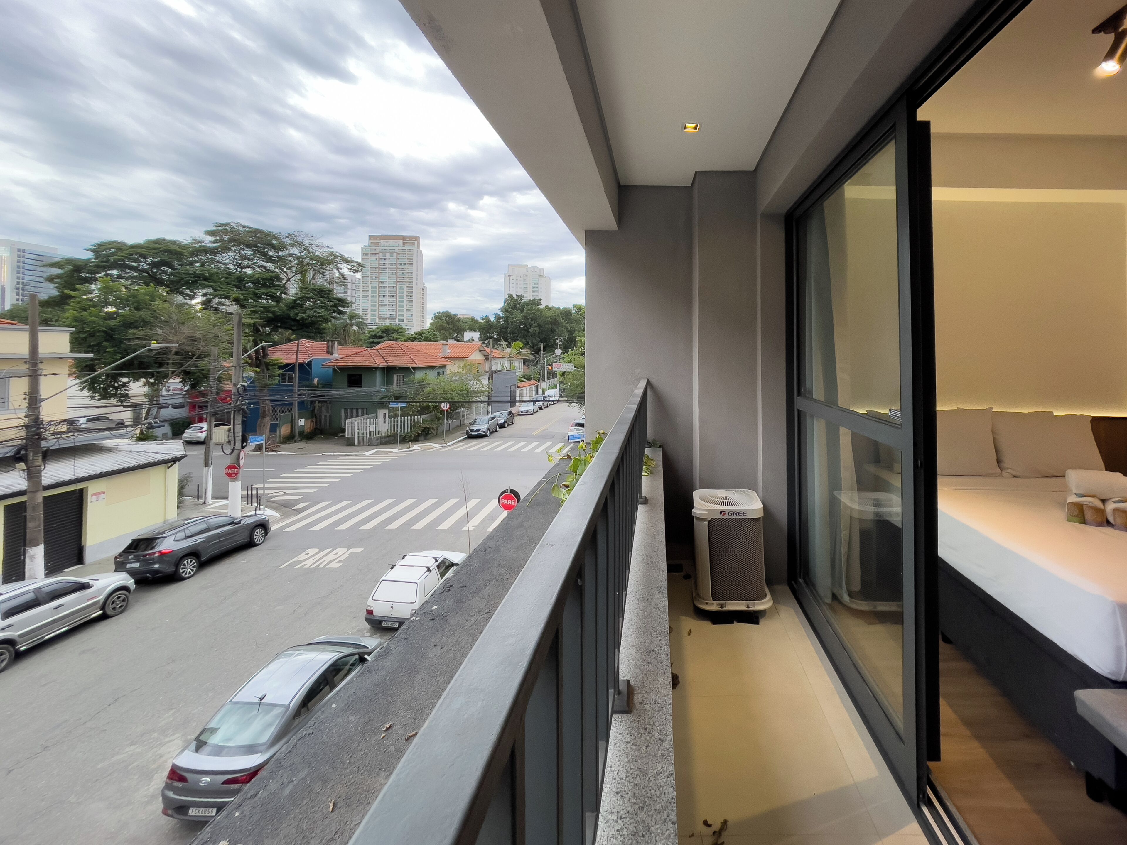 A balcony with a view of a city street, a GREE air purifier, and a bed with a white mattress and pillows.