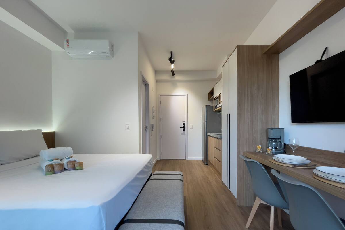 Habitación de hotel compacta con cama, escritorio, silla, televisor y kitchenette.