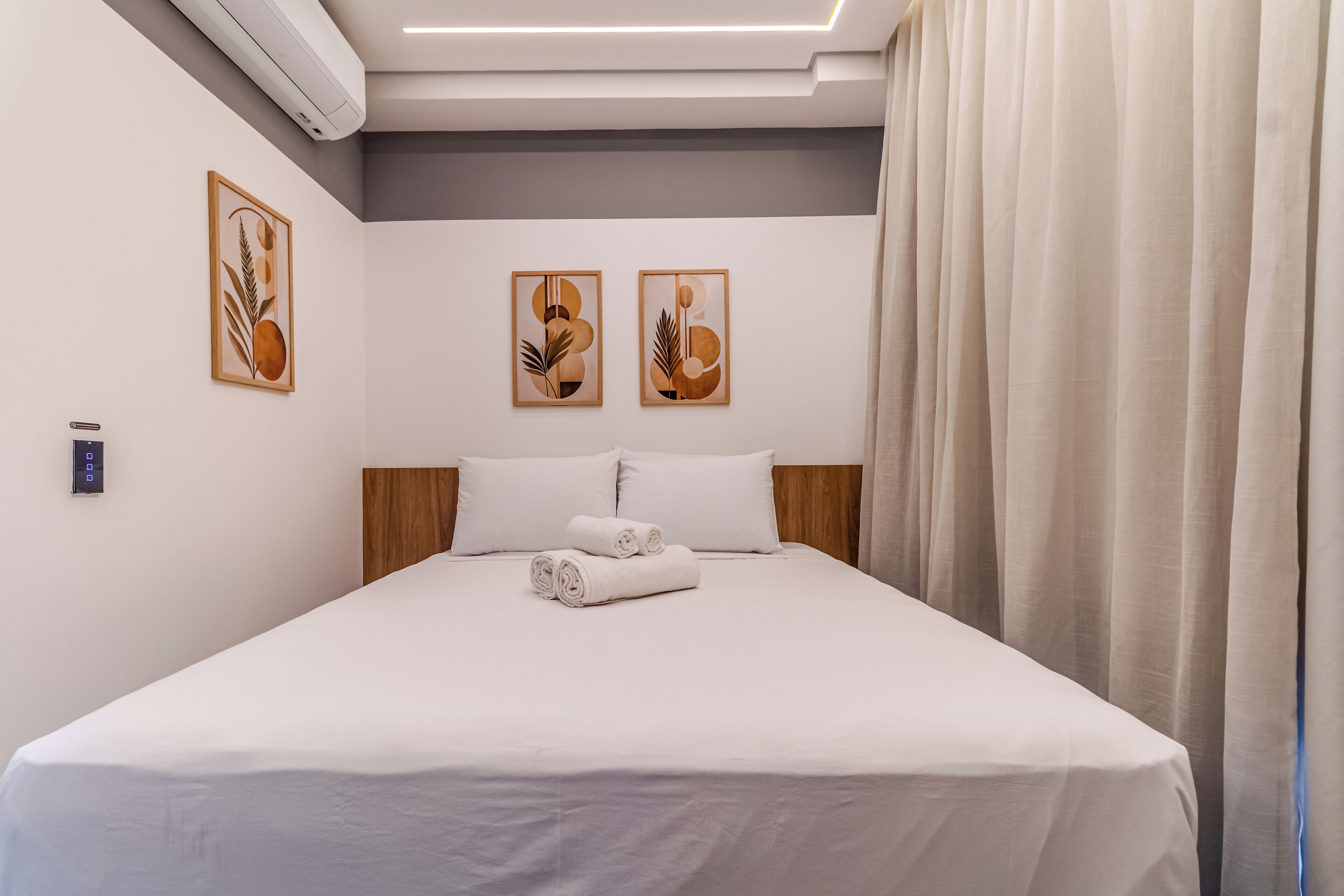 A neatly made bed with white linens and rolled towels, a wooden headboard, and two abstract paintings on the wall.