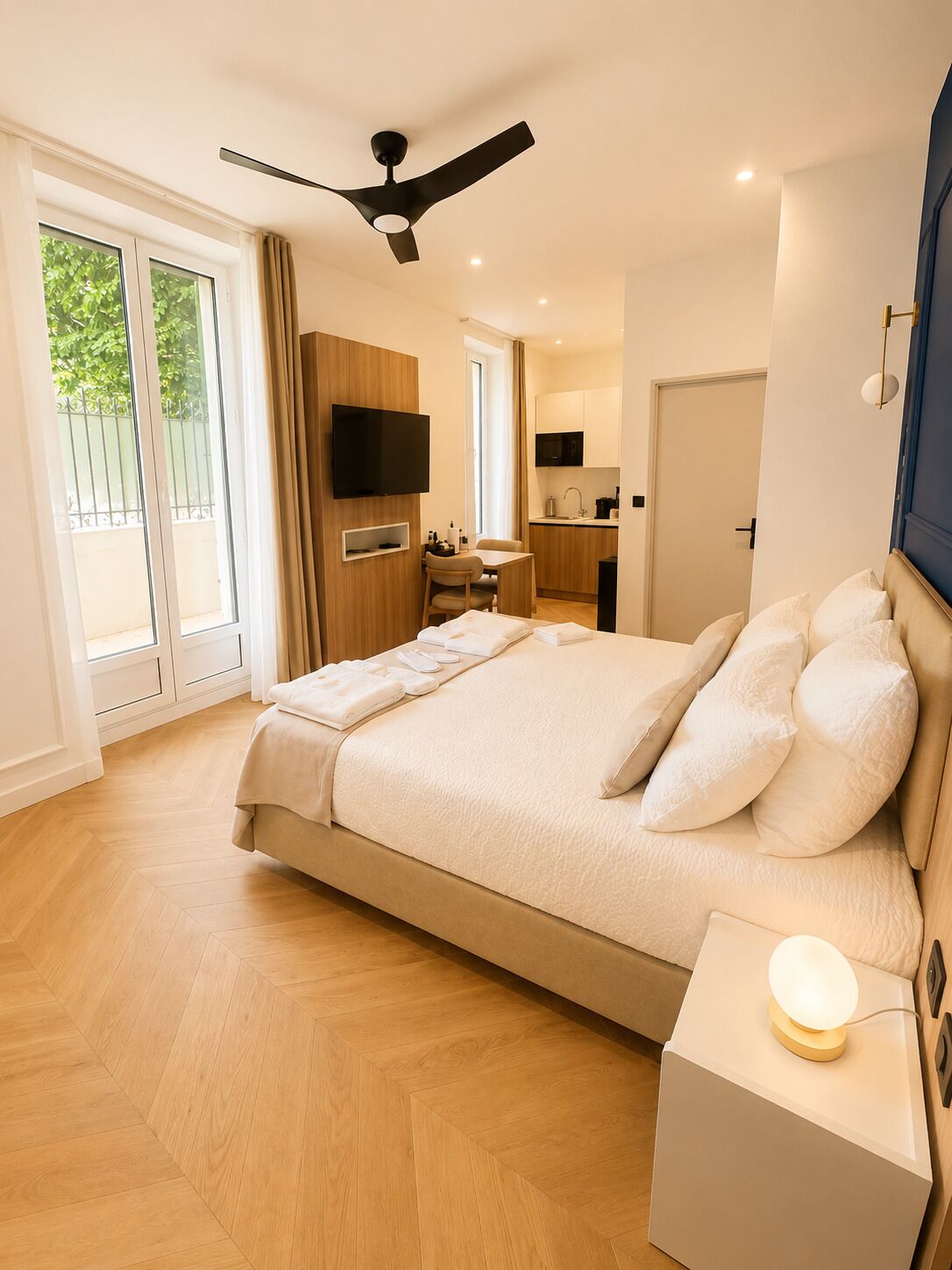A modern hotel room with a large bed, a flat-screen TV, a ceiling fan, and a small kitchenette.