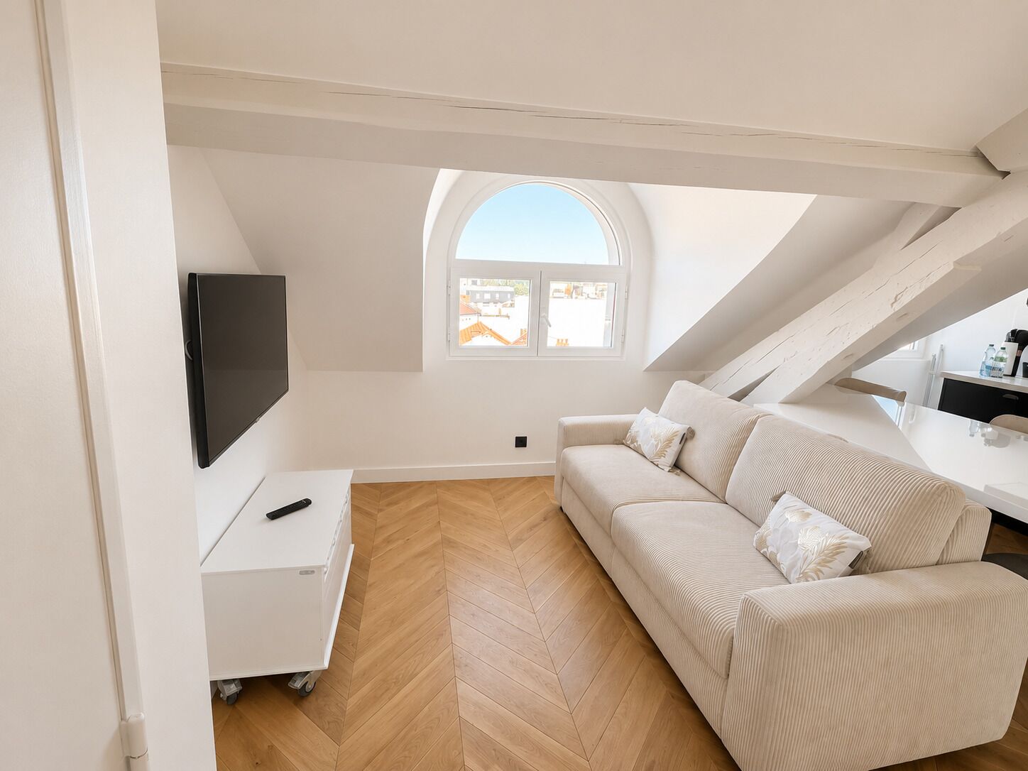 Une chambre de style loft moderne, avec un parquet en bois disposé en chevrons, un canapé blanc, un téléviseur fixé au mur et une fenêtre de toit.