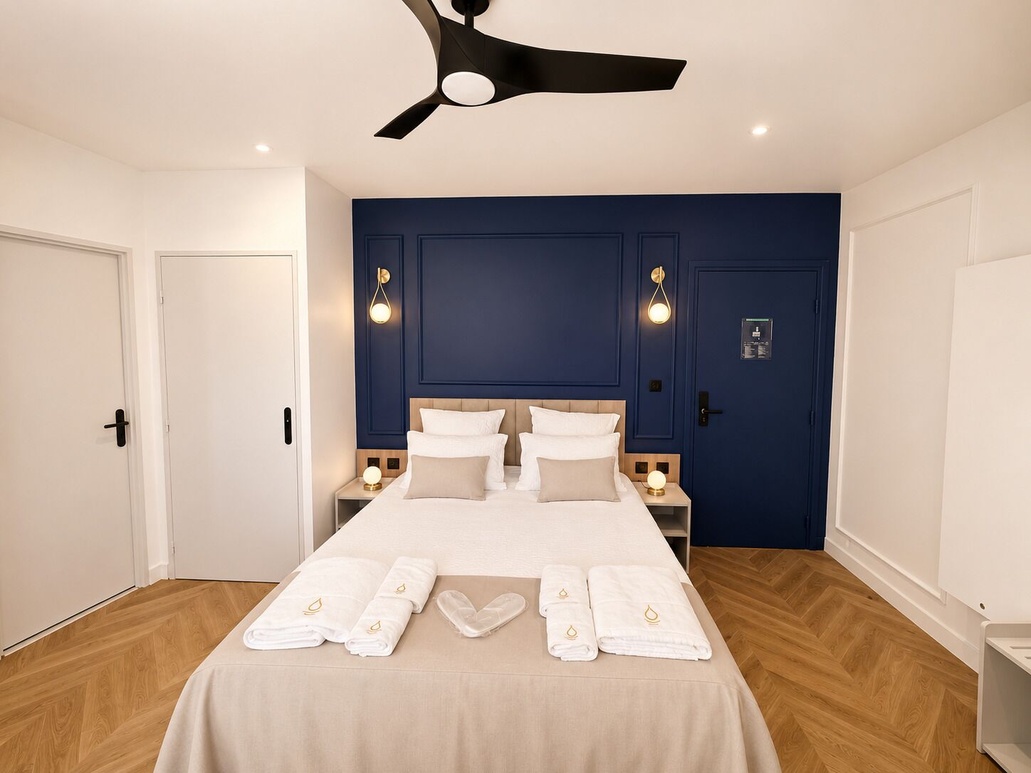 A hotel room with a bed, two bedside tables, a ceiling fan, and a blue accent wall.