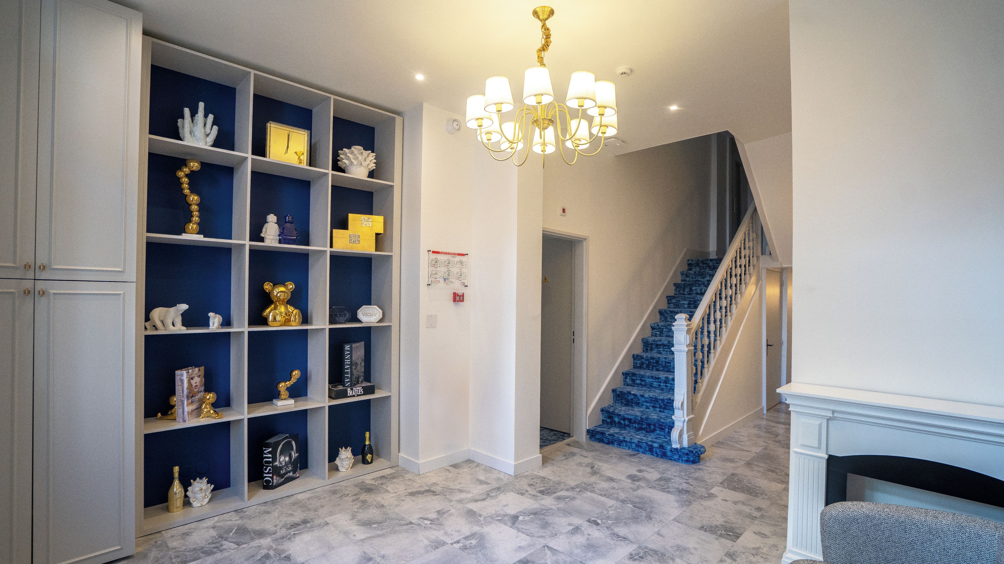 Un couloir avec un tapis bleu, des murs blancs, un lustre, un escalier aux marches bleues et une cheminée.