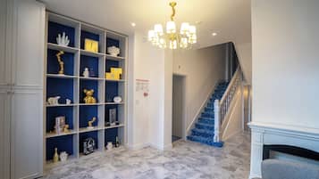 Un couloir avec un tapis bleu, des murs blancs, un lustre, un escalier aux marches bleues et une cheminée.