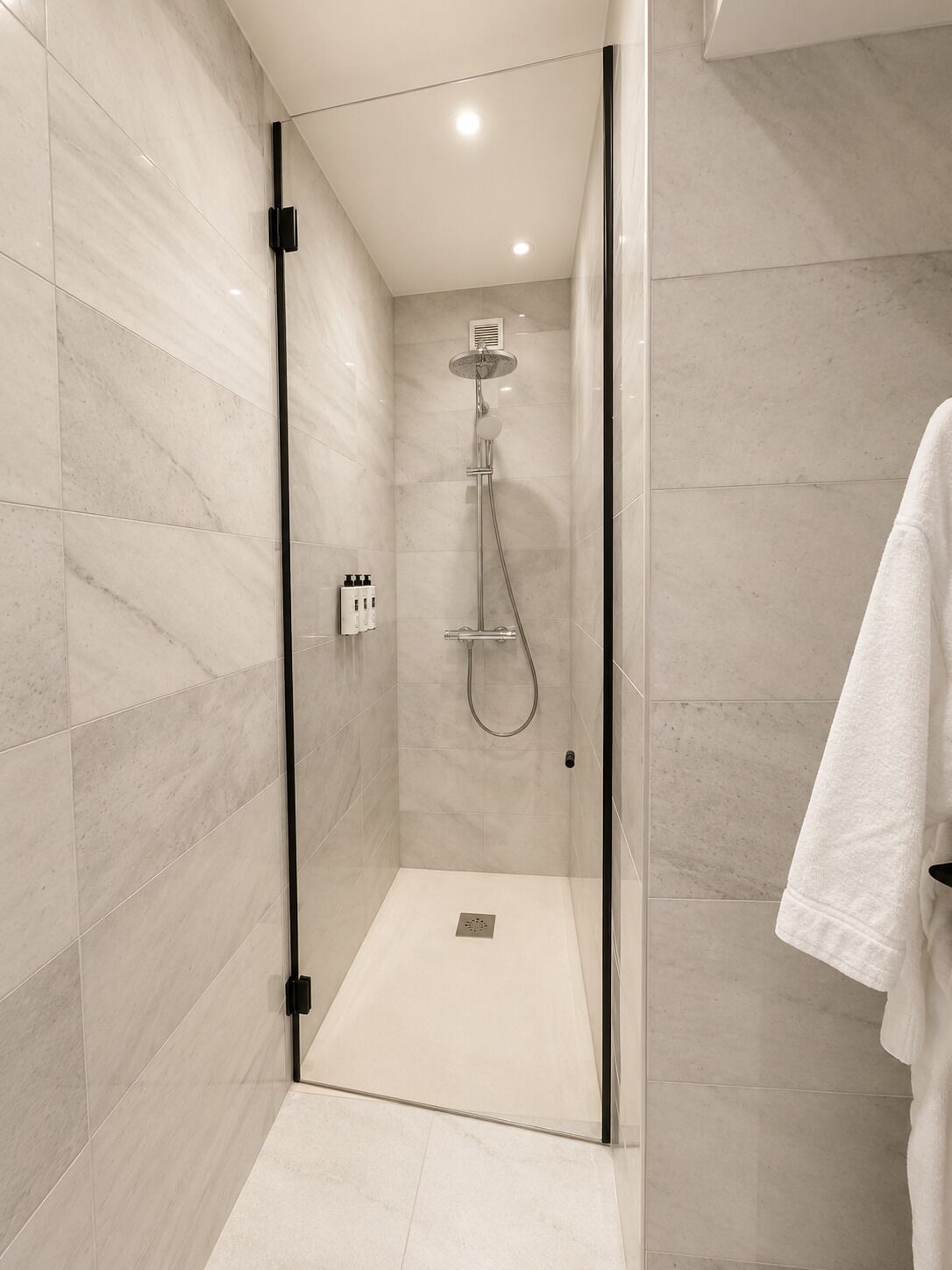 A modern shower with a glass door, a white towel hanging on the wall, and a tiled floor.
