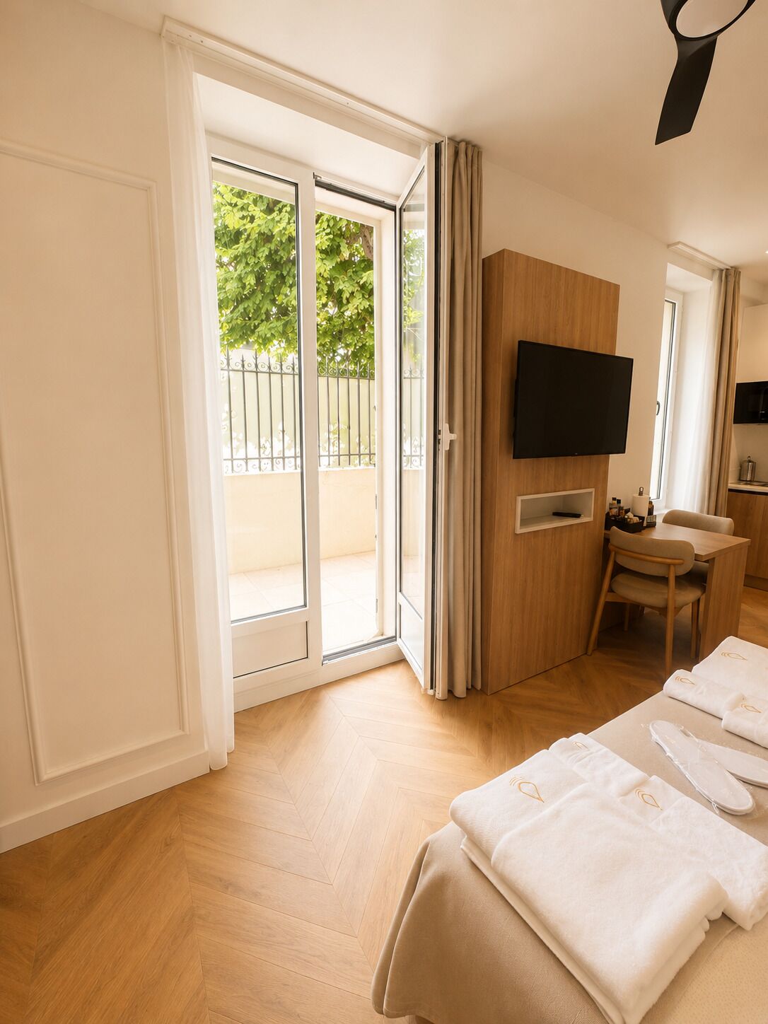 Une chambre d’hôtel avec un plancher en bois, un lit recouvert de draps blancs, un téléviseur fixé au mur et une porte-fenêtre coulissante donnant sur un espace extérieur.