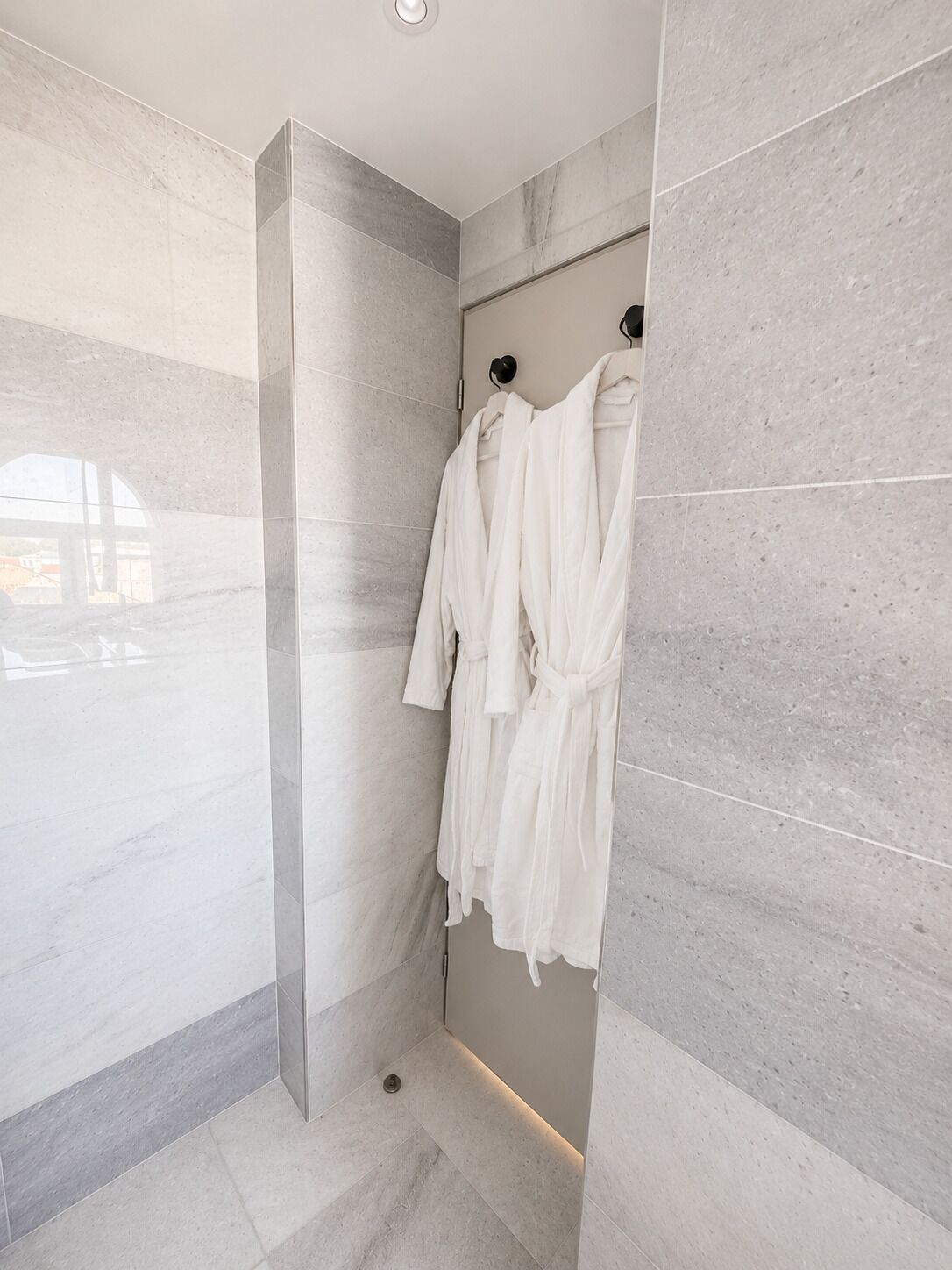 A modern bathroom with a walk-in shower, grey marble tiles, and a towel rack with white bathrobes.