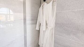 A modern bathroom with a walk-in shower, grey marble tiles, and a towel rack with white bathrobes.
