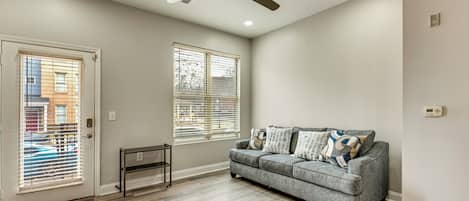 Un salon avec un canapé gris, deux fenêtres équipées de stores, un ventilateur de plafond et un plancher en bois.