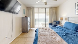A hotel room with a bed, a TV mounted on the wall, a ceiling fan, a nightstand with a lamp, and a view of the outdoors through a sliding door. - 10th-floor 3BR Beachfront Condo w/ Views & Pool (South Padre Island)