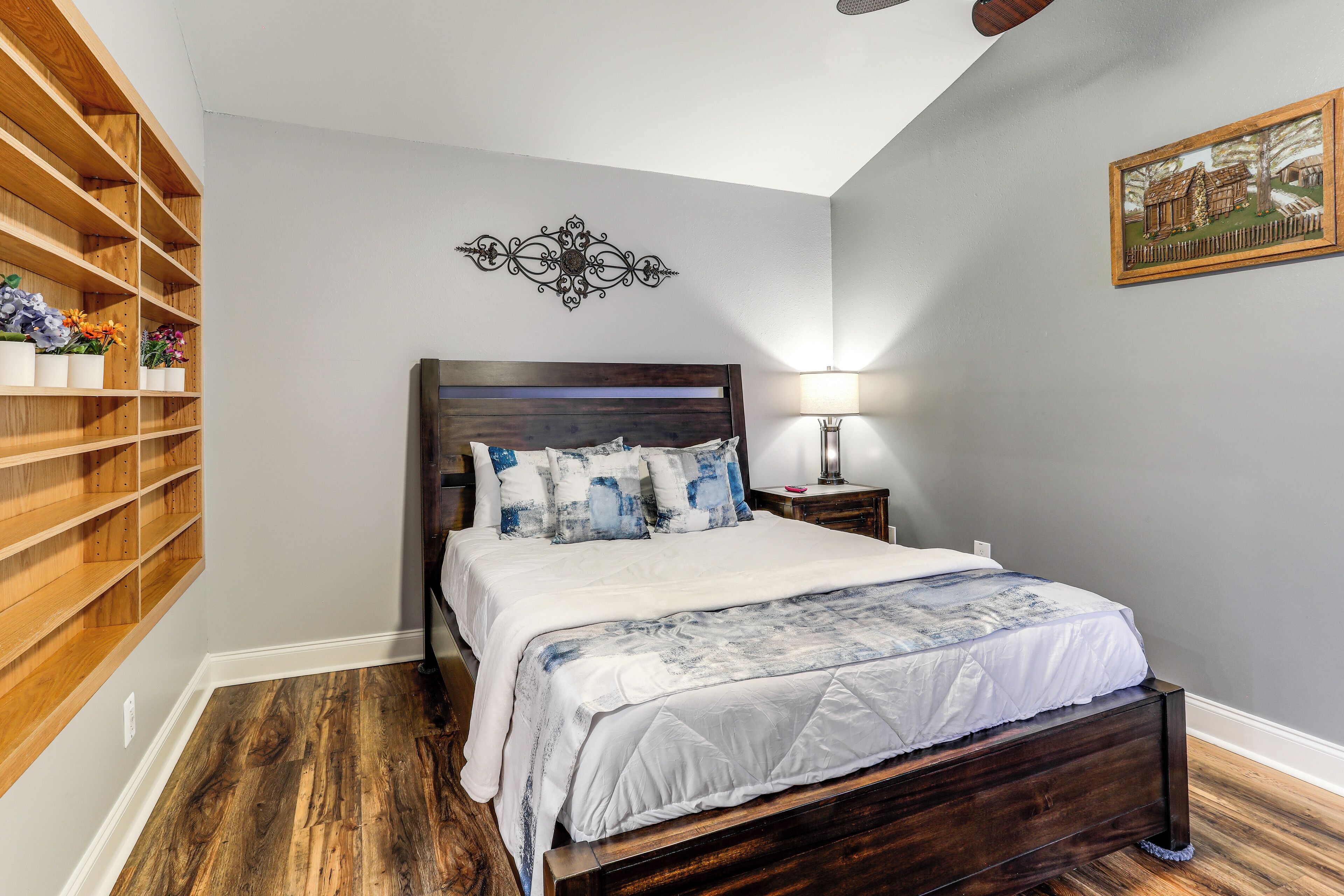 A bedroom with a wooden headboard, a bed with white bedding, a nightstand with a lamp, and a wall-mounted shelf with flowers.