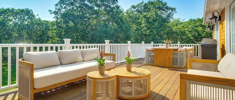 A wooden deck with a white railing, a sofa, two round tables with plants, and a grill station.