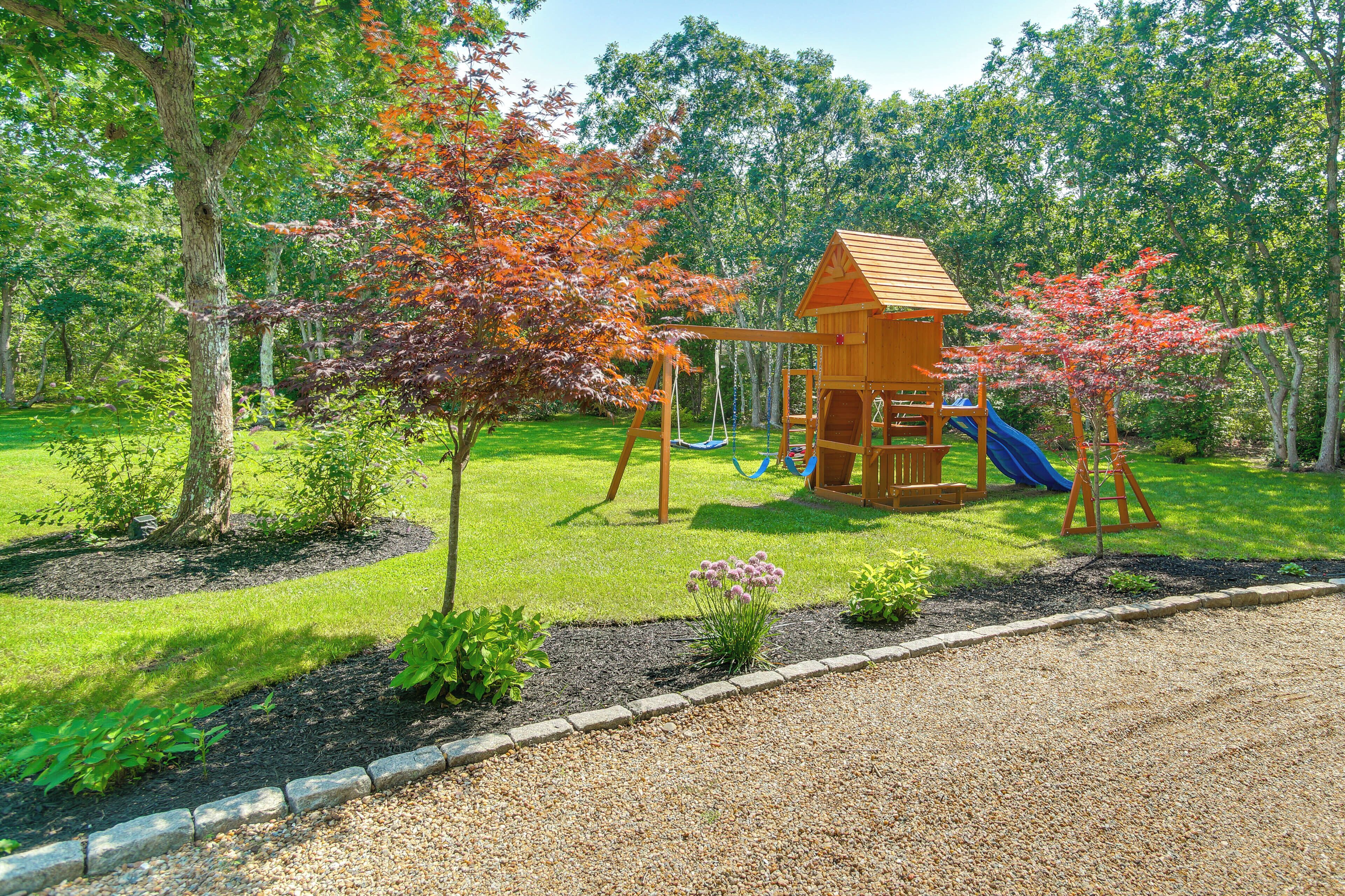 A playground with a wooden swing set and slide, surrounded by trees and a grassy area.