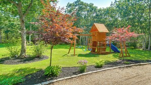 A playground with a wooden swing set and slide, surrounded by trees and a grassy area. - Summer Beach Pass: Wooded Martha's Vineyard Escape (Vineyard Haven)
