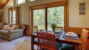 A wooden interior with a dining table set for four, a sofa, and large windows offering a forest view.