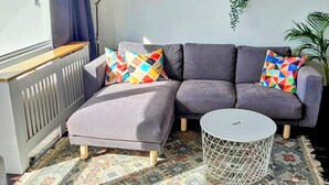 Uma sala de estar moderna com um sofá cinzento, almofadas coloridas, uma mesa de centro branca e um tapete estampado. Há quadros nas paredes, um radiador e uma janela com cortinas.