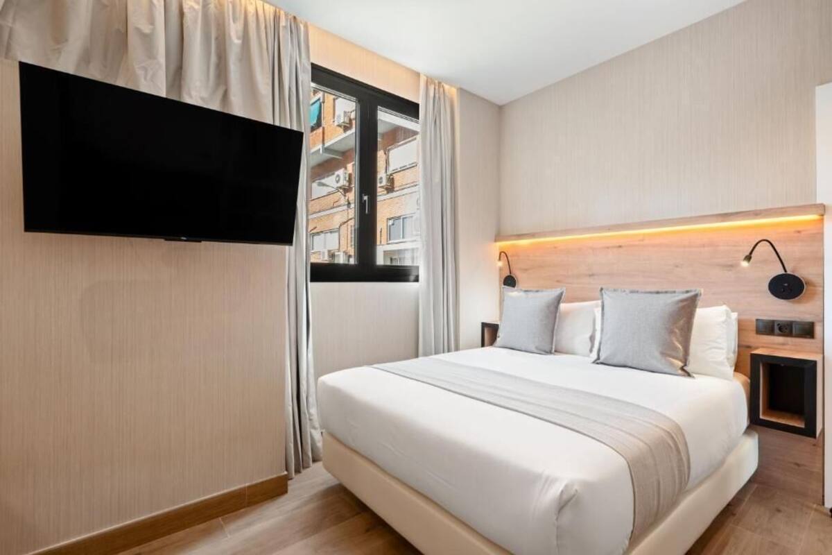 A modern hotel room with a large bed, a flat-screen TV mounted on the wall, a nightstand with a lamp, and a view of the city through a window.