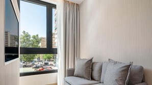 A modern living room with a grey sofa, a large window with curtains, and a view of a parking lot and buildings outside. - Dobo Madrid Betanzos (Madrid)