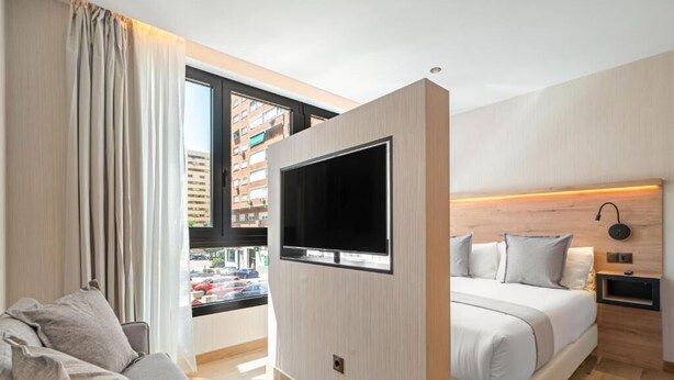 A modern hotel room with a large bed, a flat-screen TV mounted on the wall, a bedside table, and a window with a view of the city. - Dobo Madrid Betanzos (Madrid)