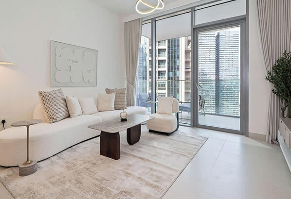 Una sala de estar moderna con un sofá blanco, una mesa de centro y un sillón. Hay una ventana grande con persianas, una planta y una obra de arte colgada en la pared.