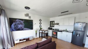 A modern living room with a large flat-screen TV, a sofa, a dining area with a table and chairs, a kitchen with white cabinets and appliances, and a decorative ceiling light. - Apartment Brisa Del Mar Punta Cana (Punta Cana)