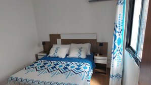 A neatly made bed with a patterned bedspread, a wooden headboard, two bedside tables with lamps, and a window with curtains. - Apartment Brisa Del Mar Punta Cana (Punta Cana)