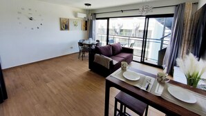 A modern living room with a wooden floor, a dining area with a table set for two, and a balcony with a view of the city. - Apartment Brisa Del Mar Punta Cana (Punta Cana)
