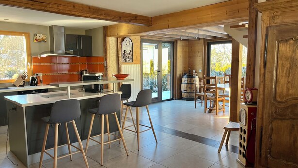 A modern kitchen with a central island, bar stools, and a dining area with wooden chairs. - Mountain vacation home 10 people village center (Prémanon)