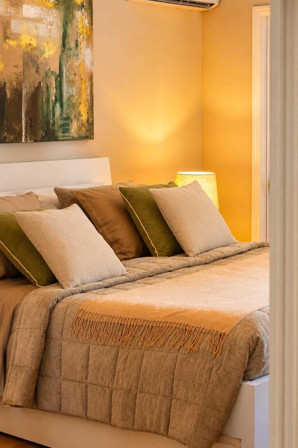 Ein ordentlich bezogenes Bett mit einer gesteppten Decke, mehreren Kissen, einer Nachttischlampe und einem Bild an der Wand.
