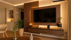 A modern living room with a flat-screen TV mounted on a wooden panel, a dining table with a Buddha statue, and a potted plant. - Luxury Apartment in Cali's Zona Rosa—Terrazas de Granada (Cali)