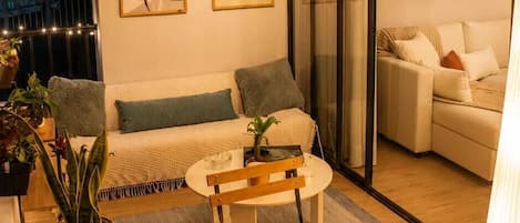 A cozy living room with a sofa, a coffee table, and a dining table. There are potted plants and artwork on the walls.