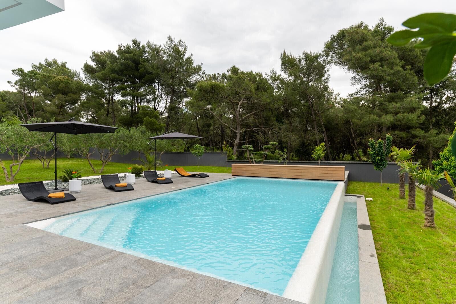 Une piscine rectangulaire avec une terrasse en bois, entourée d’une pelouse et d’arbres. Des chaises longues et des parasols sont disposés le long du bord de la piscine.