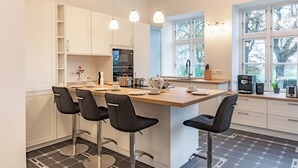 Una cucina moderna con isola centrale, sgabelli da bar, mobili bianchi e vista su alberi attraverso ampie finestre.