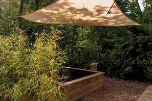 A wooden planter box with a built-in bench, surrounded by lush greenery and a large shade sail overhead.