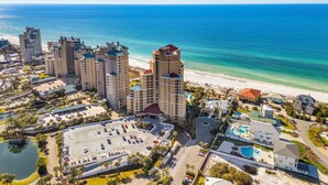 A coastal hotel complex with multiple high-rise buildings, a parking lot, and a beach. - 3BR Beachfront Resort Condo - Wraparound Balcony, Pool, Hot Tub, Tram, Beach. (Miramar Beach)