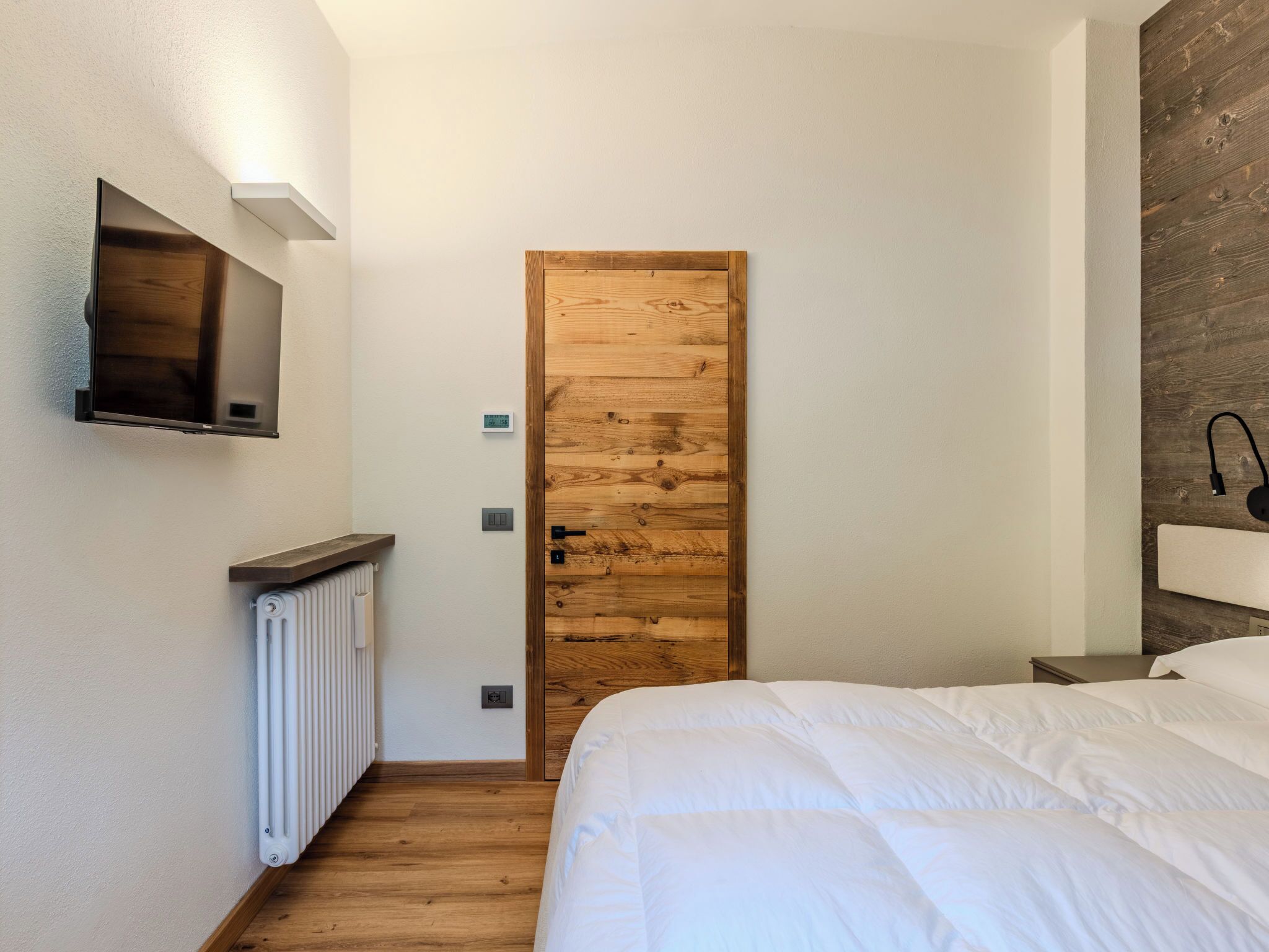 A bedroom with a wooden door, a bed, a TV mounted on the wall, a radiator, and a bedside table with a lamp.