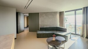 Een moderne woonkamer met een grijze hoekbank, een klein rond tafeltje en een groot raam met uitzicht op de omgeving.