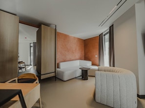 A modern hotel room with a white sofa, a round side table, a large window with curtains, and a wooden partition. - Zins Luxury Room 01 in Durr s (Durrës)