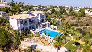 Eine großzügige, weiße Villa mit Swimmingpool, mehreren Balkonen und üppiger Bepflanzung.