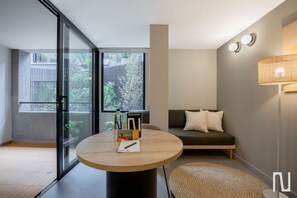 A modern living room with a round wooden table, a sofa with cushions, a large window with a view of greenery, and a wall-mounted lamp. - NOK Luxury 3BR in Zona Rosa (Bogotá)
