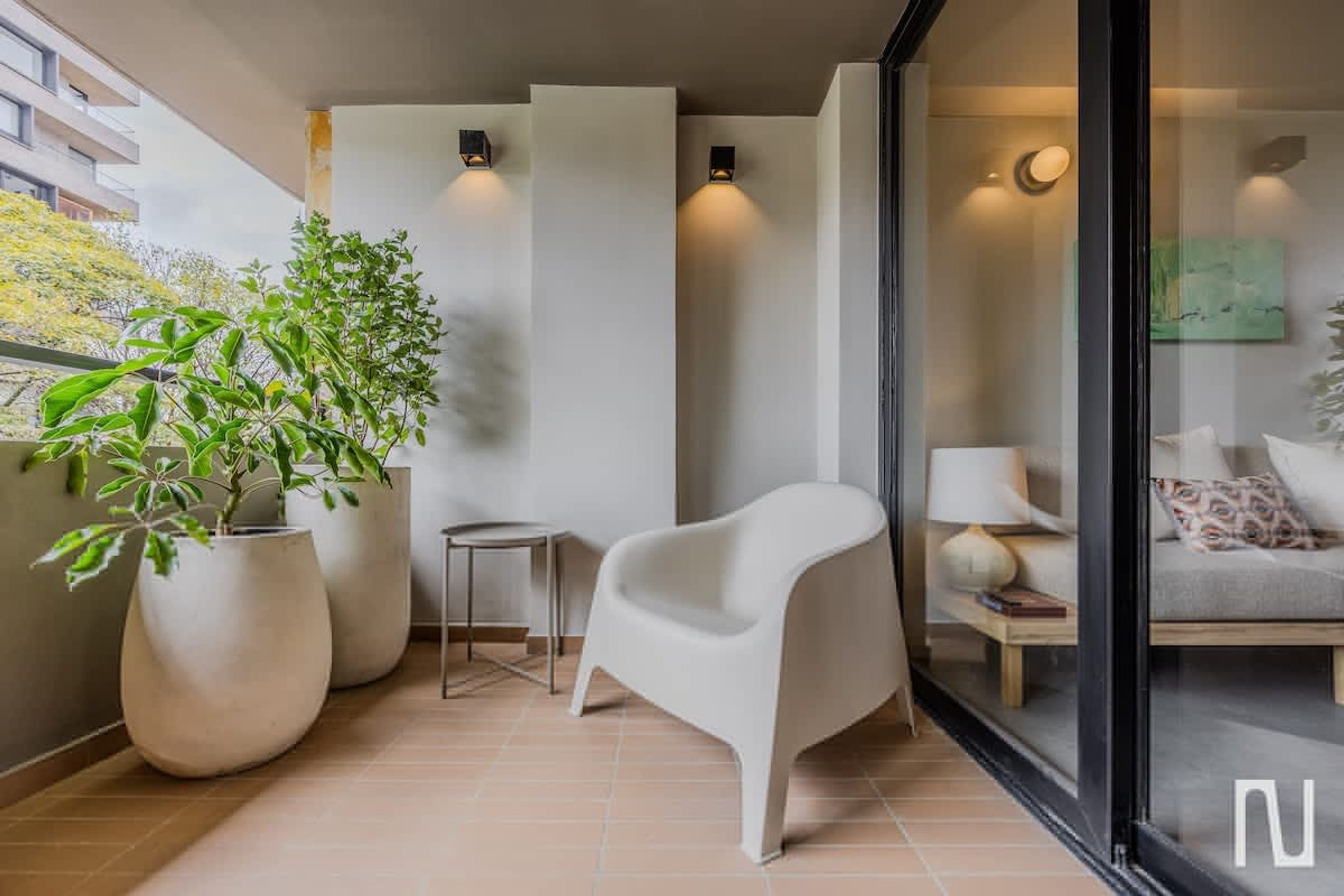 A modern balcony with potted plants, a white chair, and a glass door leading to a living area with a sofa and lamp.