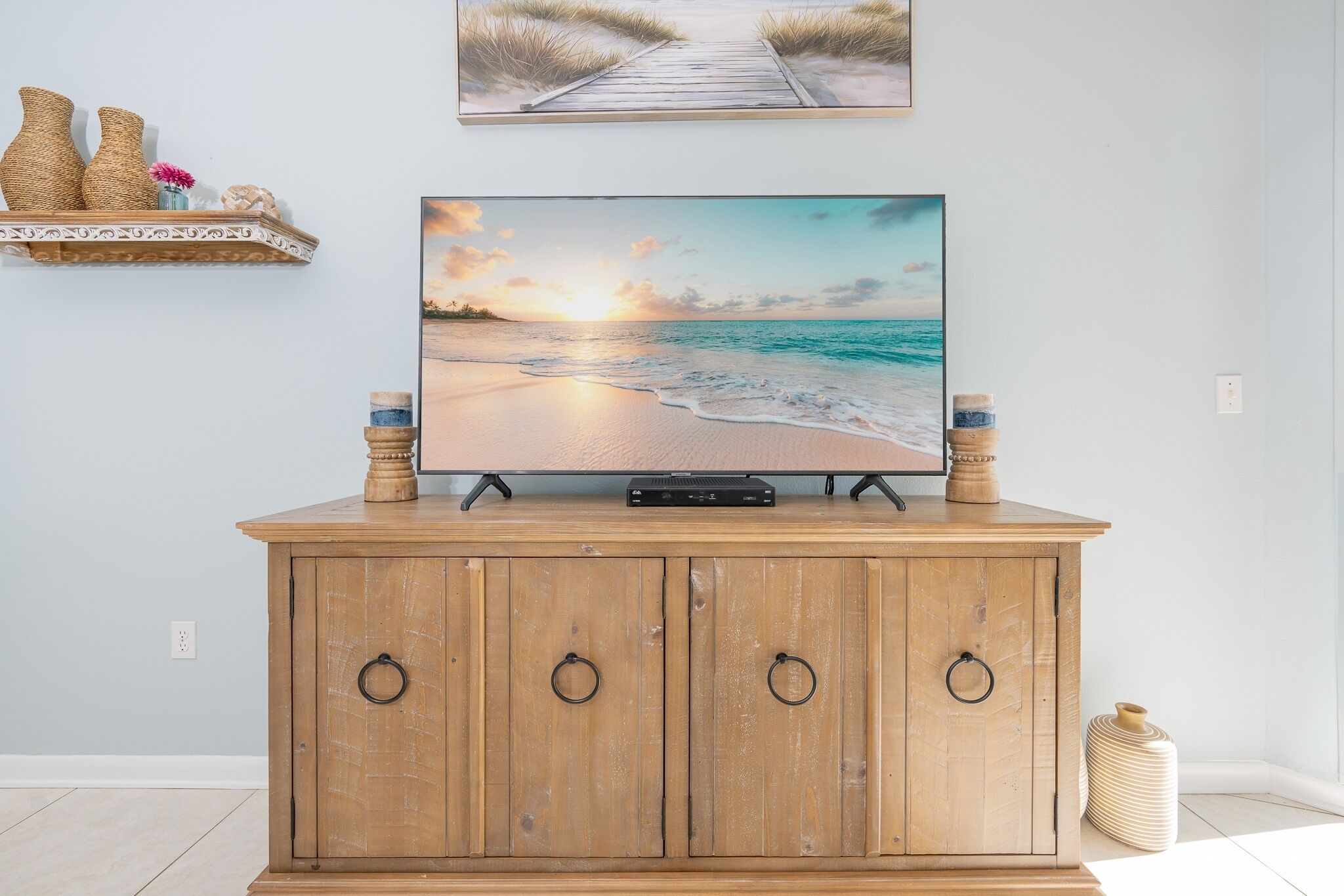 A wooden cabinet with a flat-screen TV, a shelf with decorative items, and a painting of a beach scene.