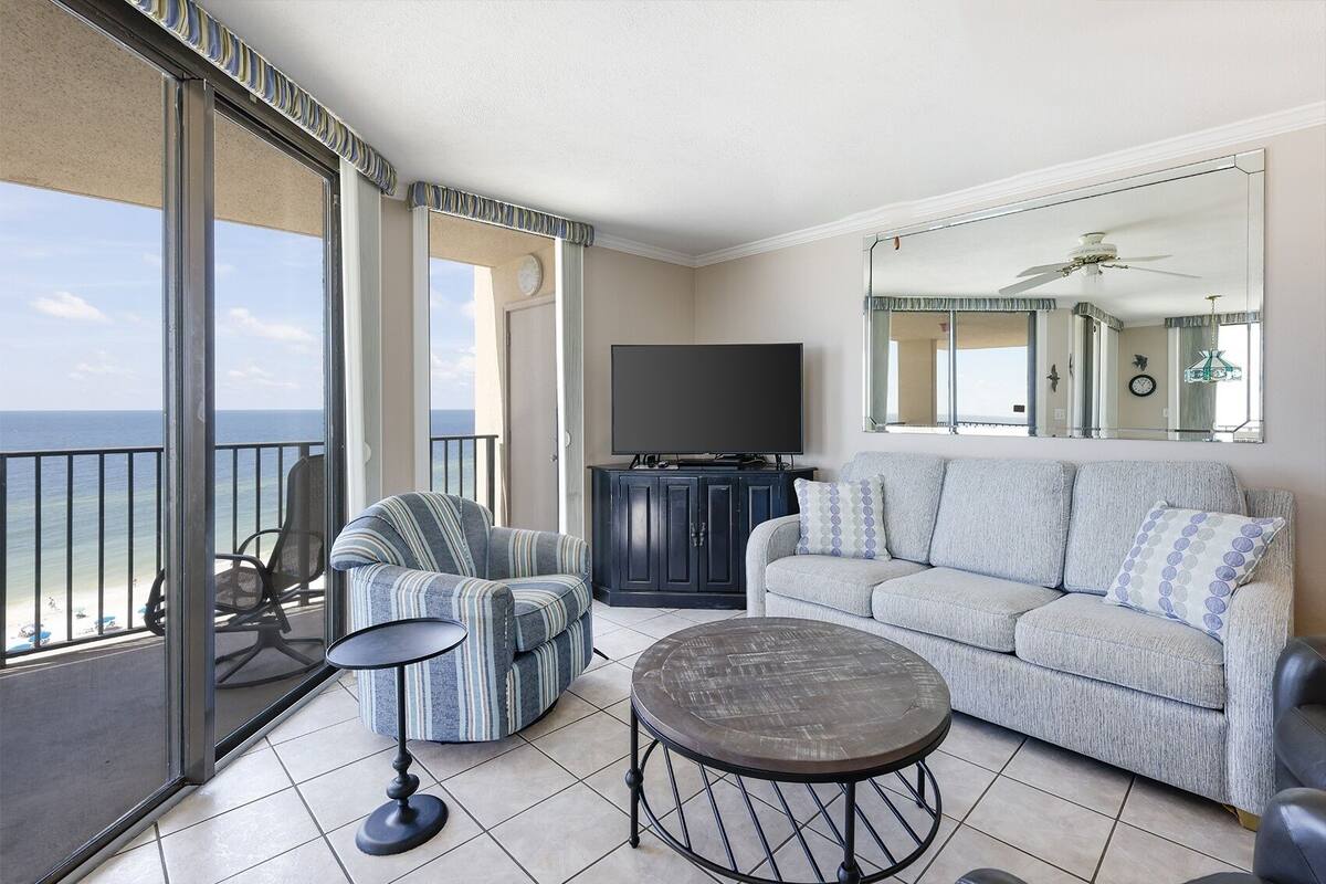 Un balcón con vistas al océano, un sofá, una silla, un mueble de televisión y una mesa redonda.